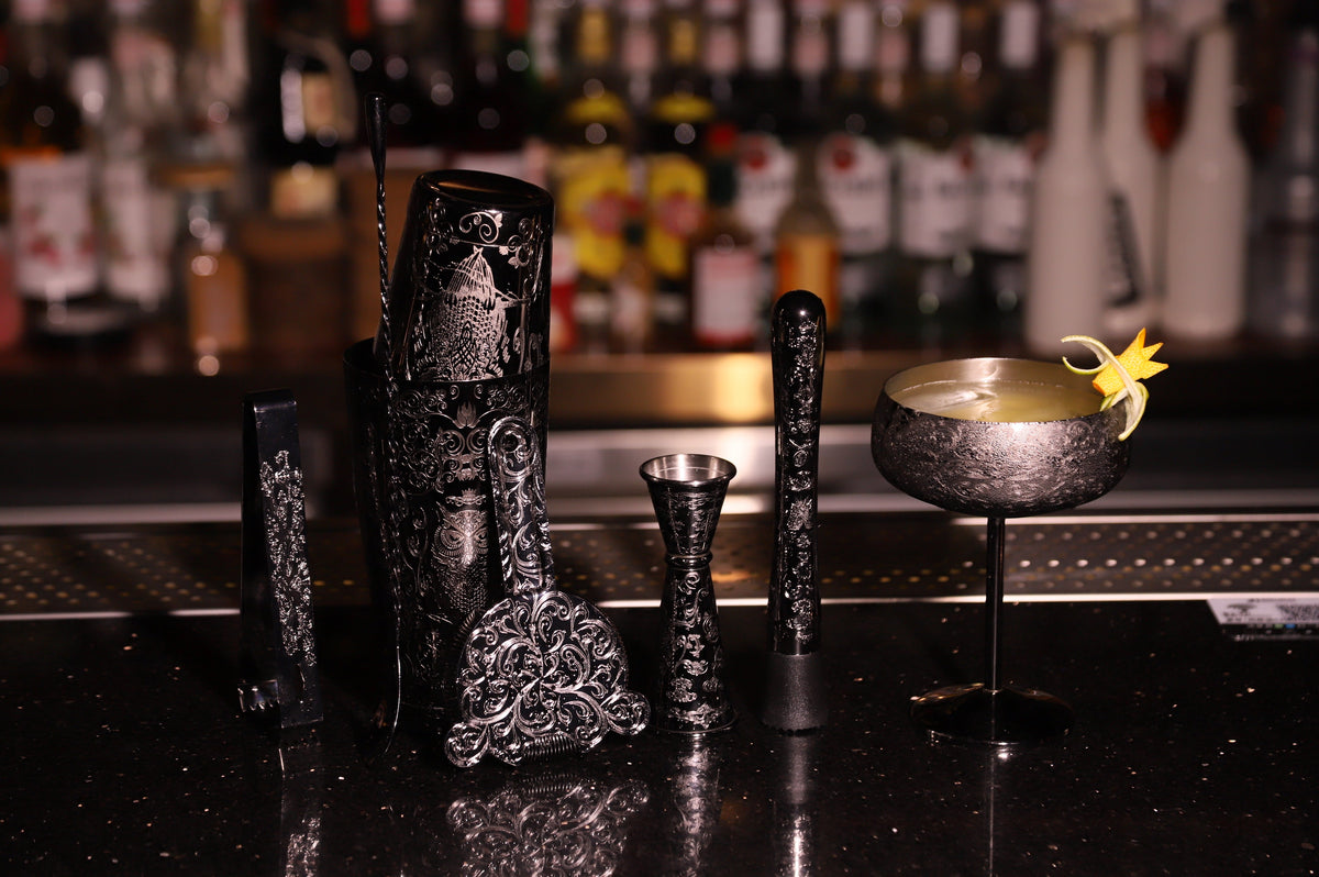 Etched black cocktail shaker set with matching bar tools displayed beside a garnished cocktail on a bar counter