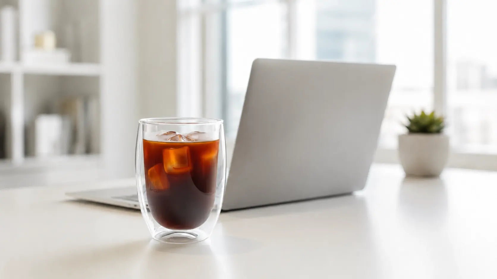 Double wall glass cup with iced coffee on a clean office desk beside a laptop