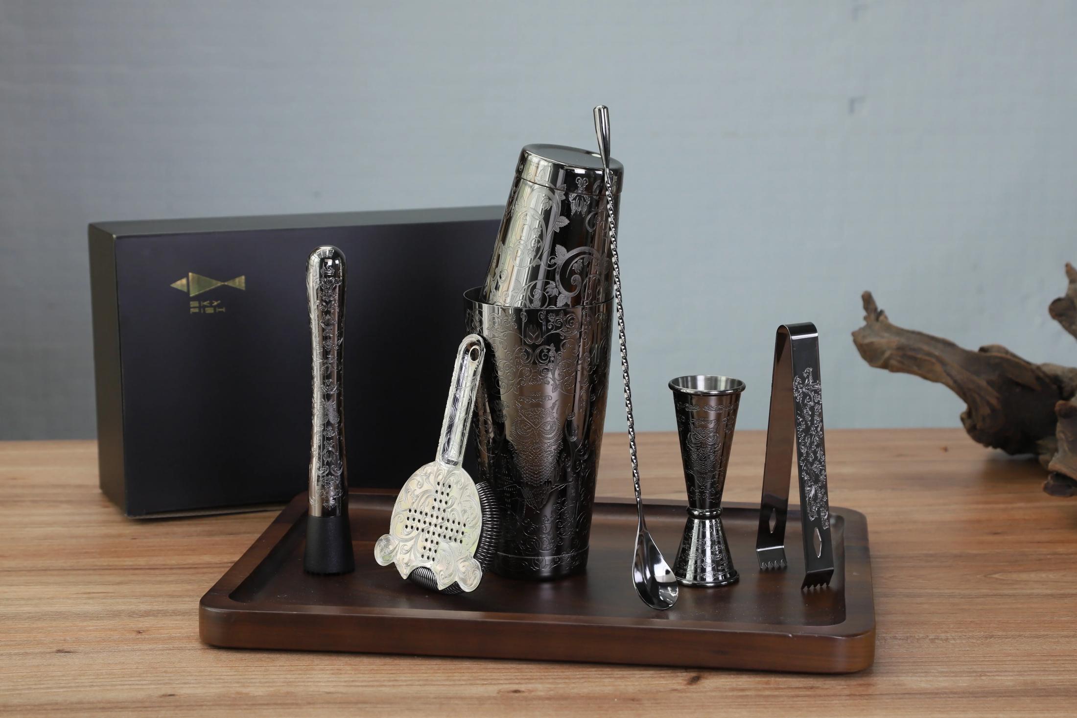 Set of cocktail tools including a shaker, jigger, and tongs on a wooden tray with a gray background.