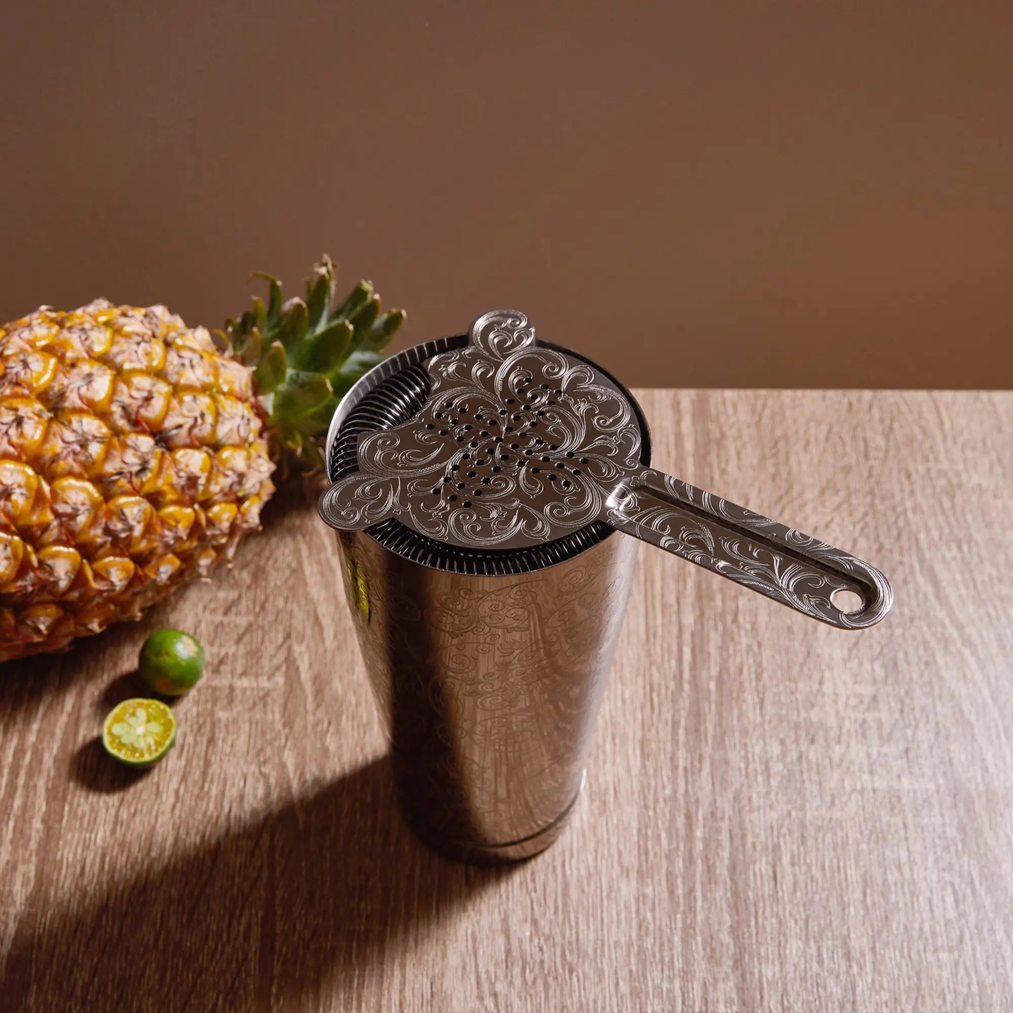 Metal cocktail shaker with decorative strainer next to a pineapple and limes on a wooden surface.