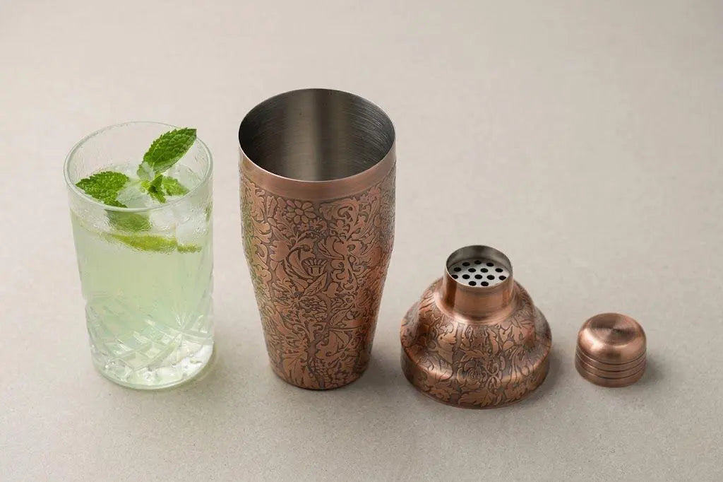 An antique copper plated 3-piece cobbler shaker disassembled into tin, built-in strainer cap, and top lid next to a cocktail glass with mint leaves.