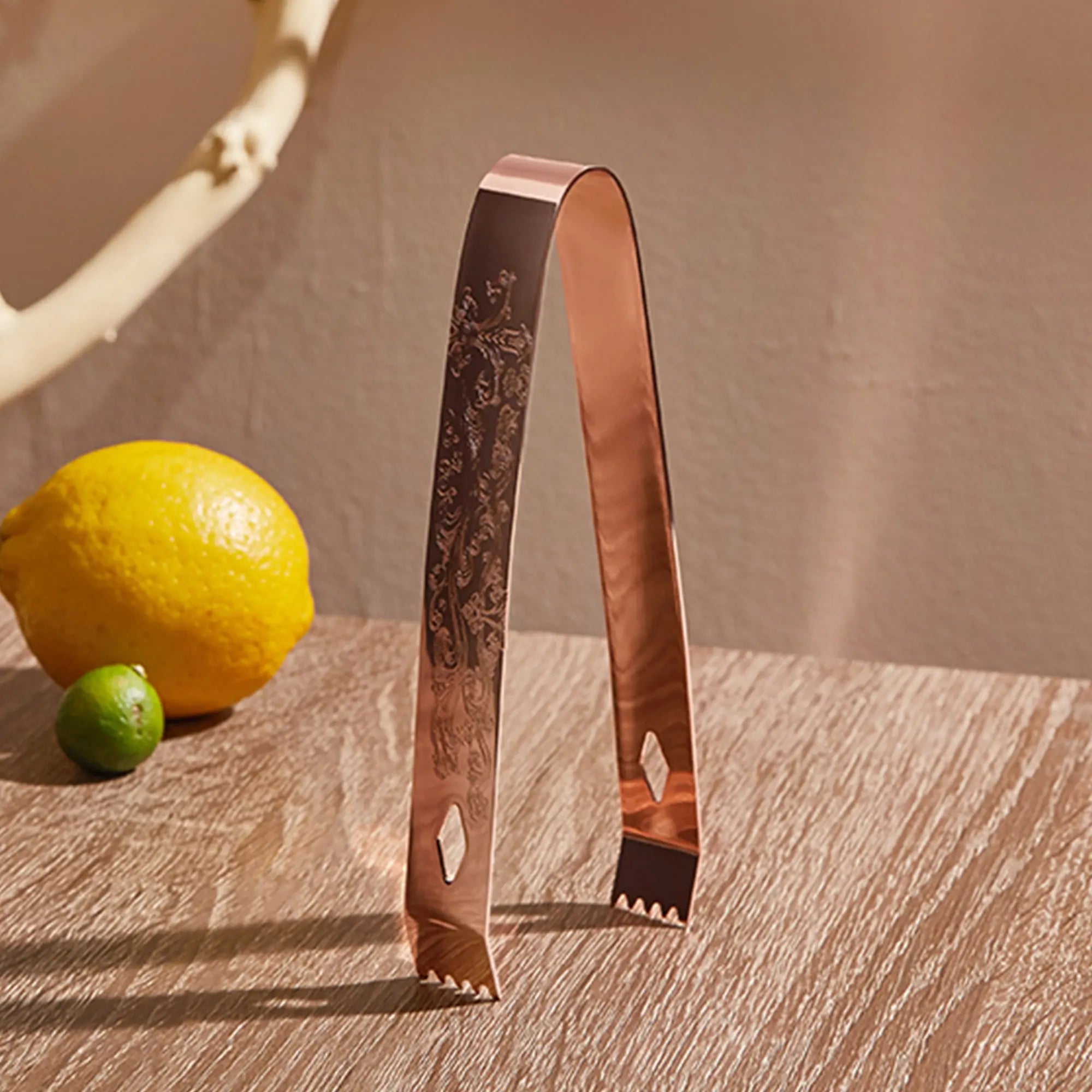 Copper tongs with intricate design on a wooden surface with a lemon and lime.
