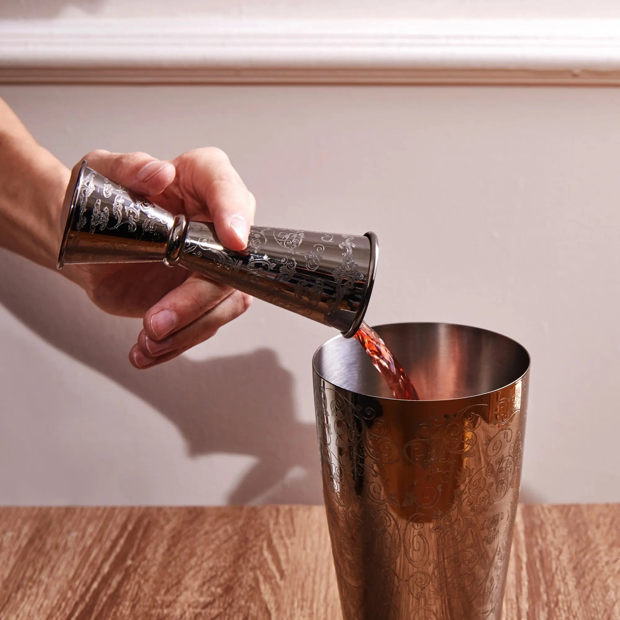 Hand pouring red liquid from a metal jigger into a metal cocktail shaker on a wooden surface.