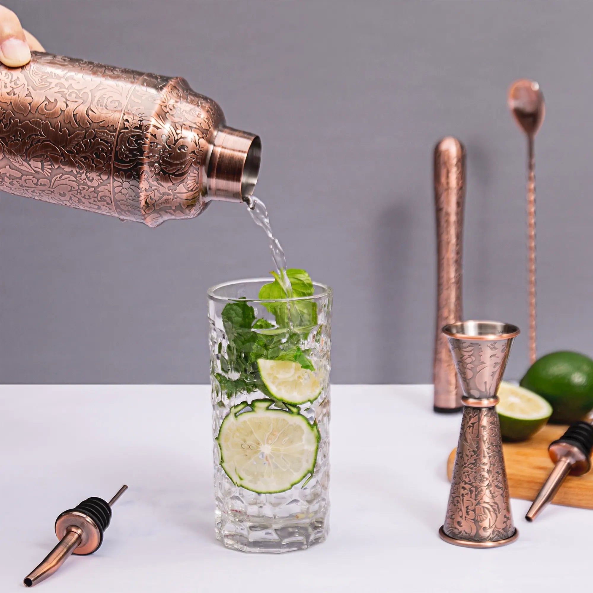 A bartender pouring a fresh mint cocktail from an antique copper plated cobbler shaker into a highball glass filled with ice and lime slices.
