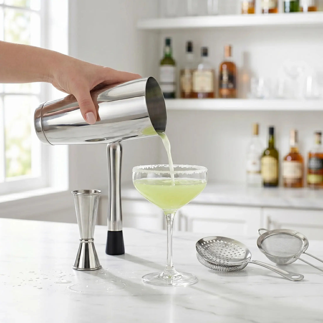 A bartender pouring a green cocktail from a polished silver stainless steel Boston shaker tin into a stemmed glass cup on a marble counter.